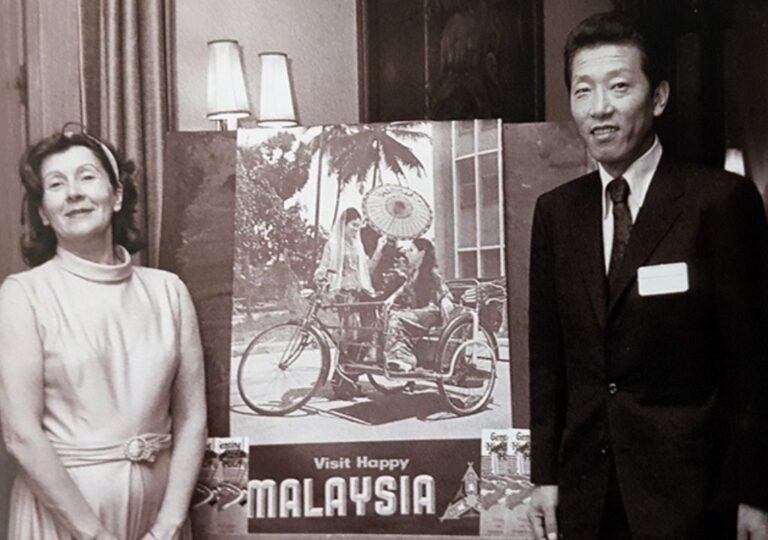 1970년대 말레이시아 관광 홍보 행사장에서 포스터 앞에 서 있는 두 사람의 흑백 기록 사진